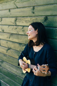 Smiling woman playing guitar while standing by wall outdoors