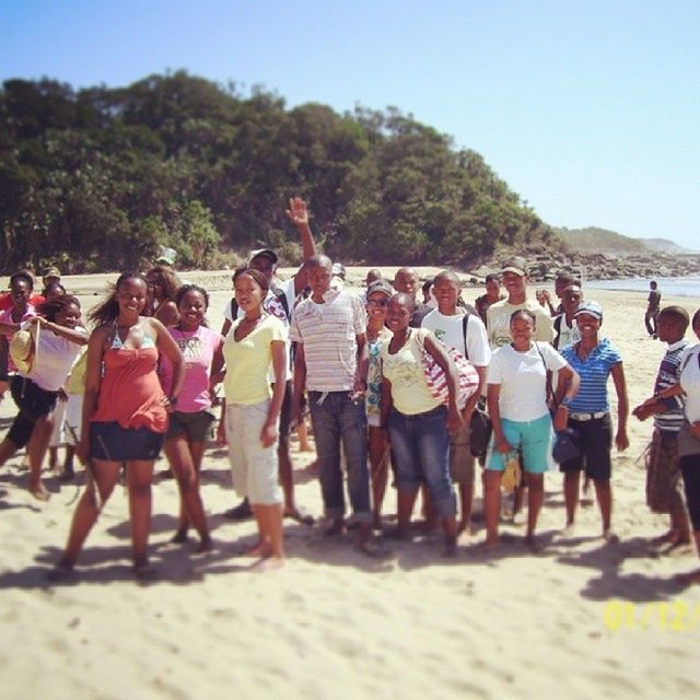 large group of people, beach, lifestyles, leisure activity, person, sand, men, togetherness, vacations, enjoyment, clear sky, tourist, shore, mixed age range, standing, casual clothing, sea, mountain, tree