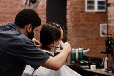 Barber in protective mask and latex glove styling adult man in a barbershop salon. new normal