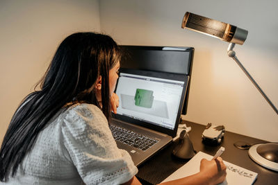 Rear view of young businesswoman sitting at desk, using laptop and 3d design software