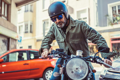Serious bearded man in sunglasses and helmet sitting on motorbike during ride on city street looking away