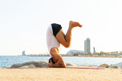 Full body unrecognizable barefoot male doing salamba sirsasana pose on beach near sea during yoga session in morning