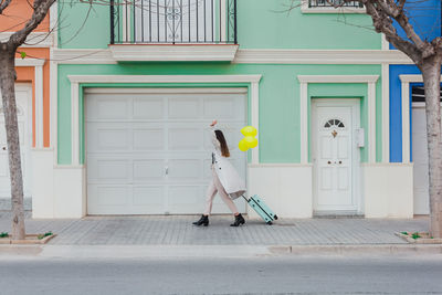 Side view of unrecognizable stylish female covering face with yellow balloons walking with suitcase on city street next to old styled colorful building