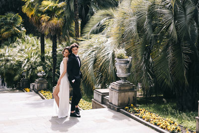 Happy lovers the bride and groom in wedding outfits walk among plants and palm trees in the old park