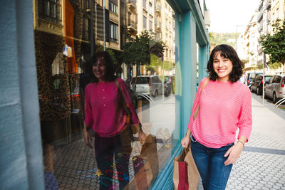 Portrait of smiling young woman holding shopping bag while walking by glass window