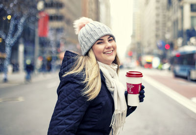 Portrait of woman holding disposable cup while standing on road in city