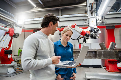 Smiling engineer standing with colleague holding laptop in robot factory