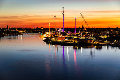 Sailboats on river by illuminated buildings against sky during sunset