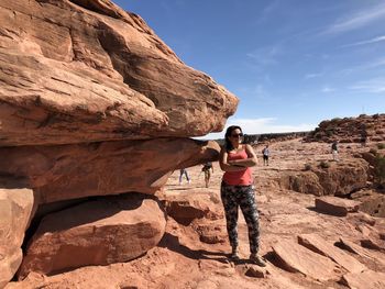 Mature woman wearing sunglasses with arms crossed standing on rock against sky during sunny day