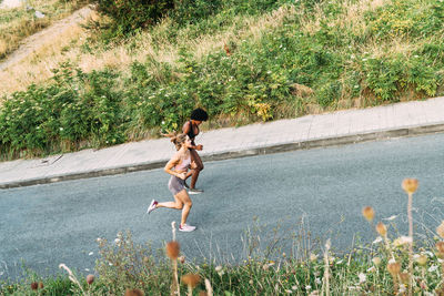 Full body of optimistic young athletic females in activewear running up asphalt road while training together near city in summer day