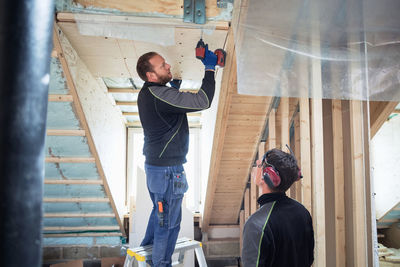 Coworker looking at carpenter drilling on roof beam in construction site