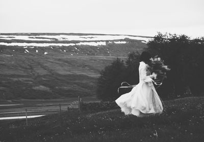 Side view of woman with bouquet walking on field against clear sky