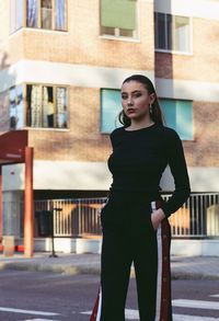 Portrait of confident young woman with hands in pockets standing against buildings in city