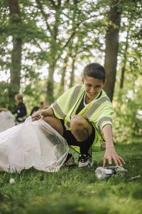 Full length of smiling boy crouching and collecting garbage in bag on grass