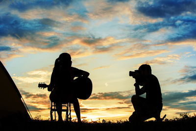 Silhouette man photographing friend playing guitar during sunset