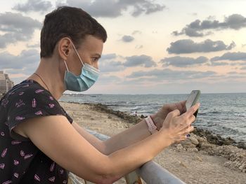 Woman wearing mask using phone standing by sea against sky