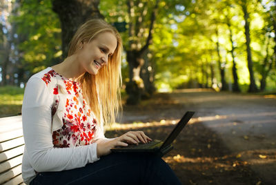 Happy beautiful woman using laptop while sitting on bench at park