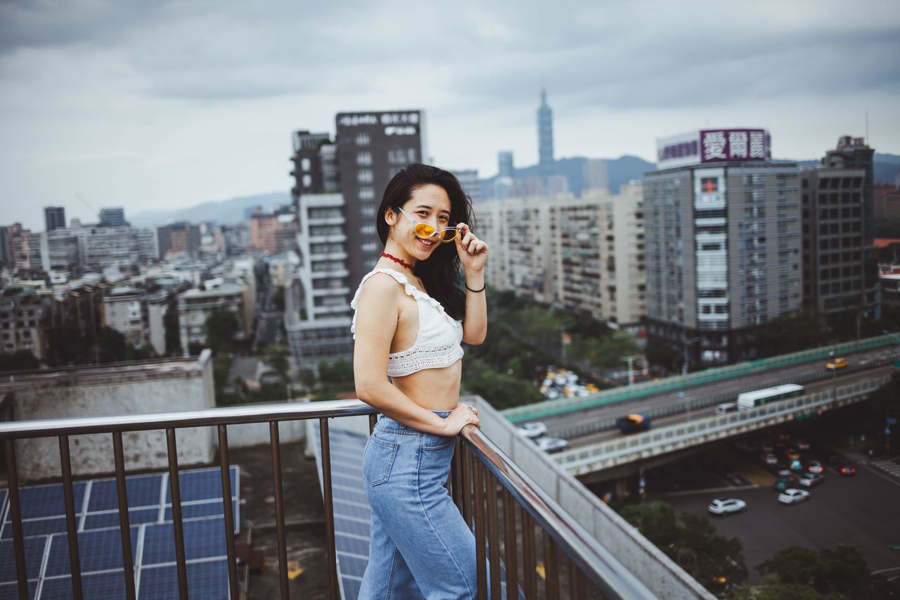 architecture, building exterior, built structure, city, standing, real people, young women, young adult, one person, casual clothing, connection, three quarter length, leisure activity, lifestyles, front view, looking at camera, railing, sky, portrait, cityscape, outdoors, beautiful woman