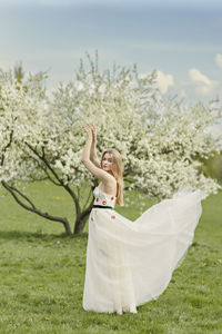 A young blonde in a long white dress poses near a cherry blossom in the garden, a spring landscape.