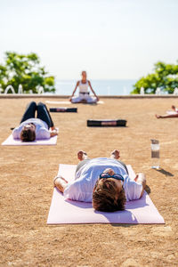 Yoga instructor in activewear sitting in lotus position while people lying on mats on ground during shavasana in park in sunny day