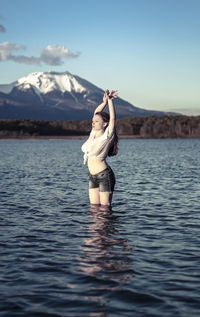 Young woman with arms raised standing in lake against snowcapped mountain