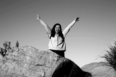 Portrait of happy woman with arms raised standing on rock against clear sky