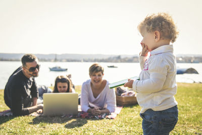 Family enjoying on grassy field by lake against clear sky during sunny day