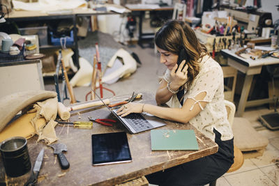 Female upholstery worker talking on phone while using laptop at workbench in workshop