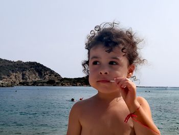 Shirtless girl eating candy at beach against clear sky