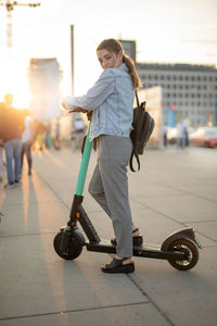 Young woman looking away while standing on push scooter against sky in city