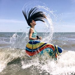Side view of girl tossing hair while standing in sea against sky during sunny day