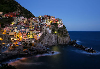 Illuminated manarola at sea shore against clear sky during dusk