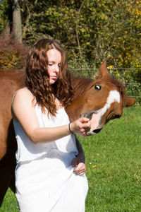 Young woman feeding horse while standing against trees during sunny day