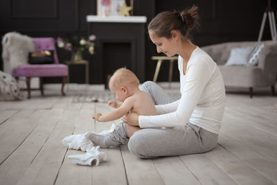 Mom changes clothes crying baby of 10 months on the floor in a cozy real light interior.