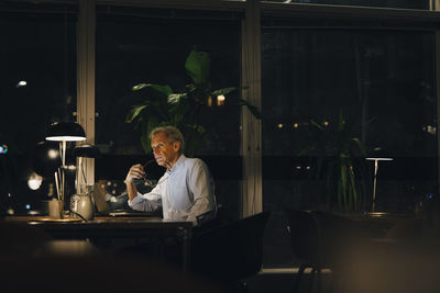 Thoughtful senior male professional looking away while sitting at desk in dark office