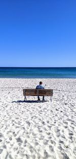 Men sitting on bench at beach against clear blue sky