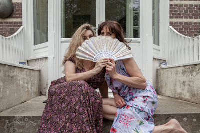 Pair of beautiful middle aged women waving a vintage fan in front of a french villa
