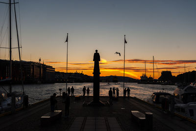 Silhouette people on street by sea against sky during sunset