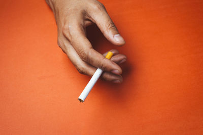 Close-up of cropped hand holding cigarette on orange background