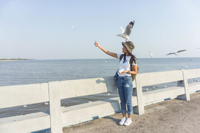 Full length of smiling woman feeding seagull standing against sea