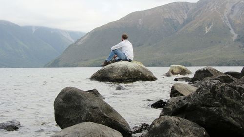 Side view of mature man sitting on rock in lake against mountain