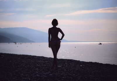 Rear view of seductive woman standing at beach against sky during sunset