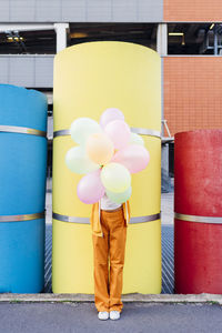 Young woman covering face with colorful balloons in front of pipes