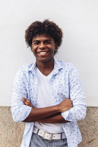 Smile african american male in trendy outfit and with afro hairstyle leaning on stone wall of building and looking at camera