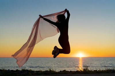Silhouette slender girl at sunset on seashore rejoices with a transparent cloth