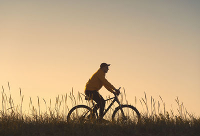 Silhouette man riding bicycle on field against sky during sunset