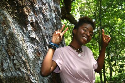 Portrait of smiling woman gesturing peace sign while standing against tree at park