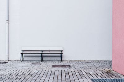 Empty bench on sidewalk against white wall