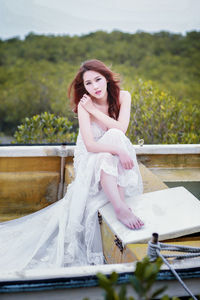 Portrait of beautiful female model in white evening gown sitting on boat against forest