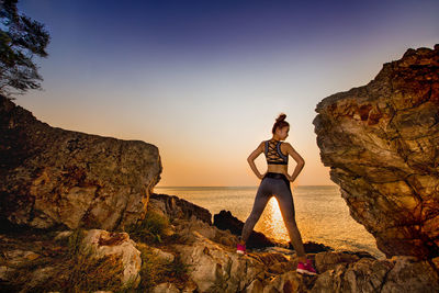 Rear view of young woman with hands on hip standing at rocky beach against clear sky during sunset